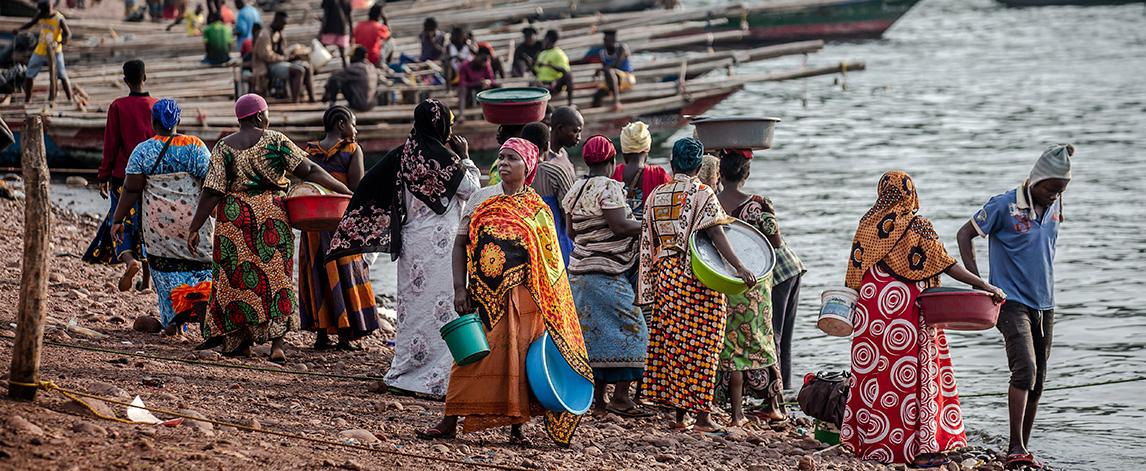 Foreground: Women gather on a beach with buckets to collect fish; background: boats lined up on shore